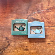 Two miniature metal watering cans in light blue and green colors, packaged in cardboard boxes with a window displaying the product. Set on a wooden surface.